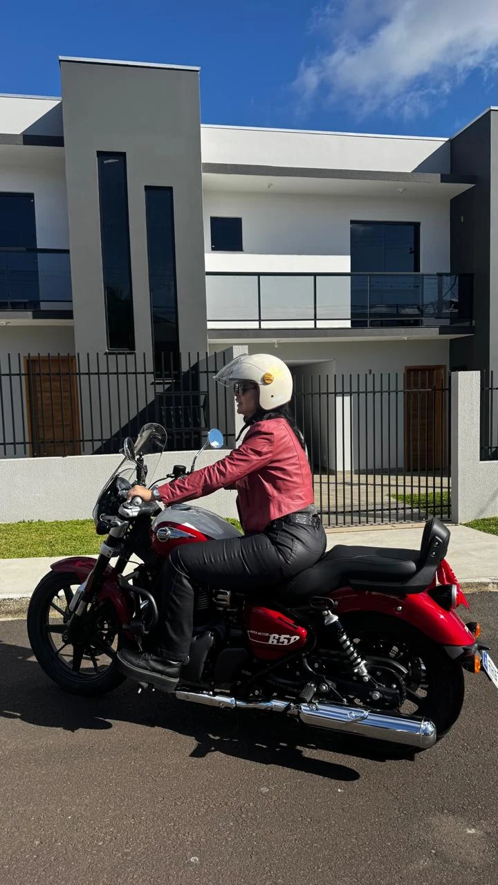 Carol de jaqueta bordô e capacete branco pilotando sua motocicleta bordô.