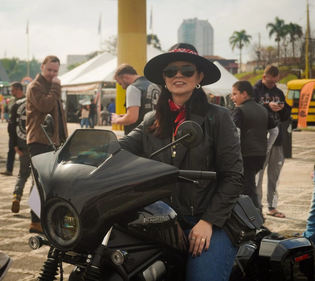 Carol com seu chapéu e bandana, sentada em uma motocicleta preta durante um evento.