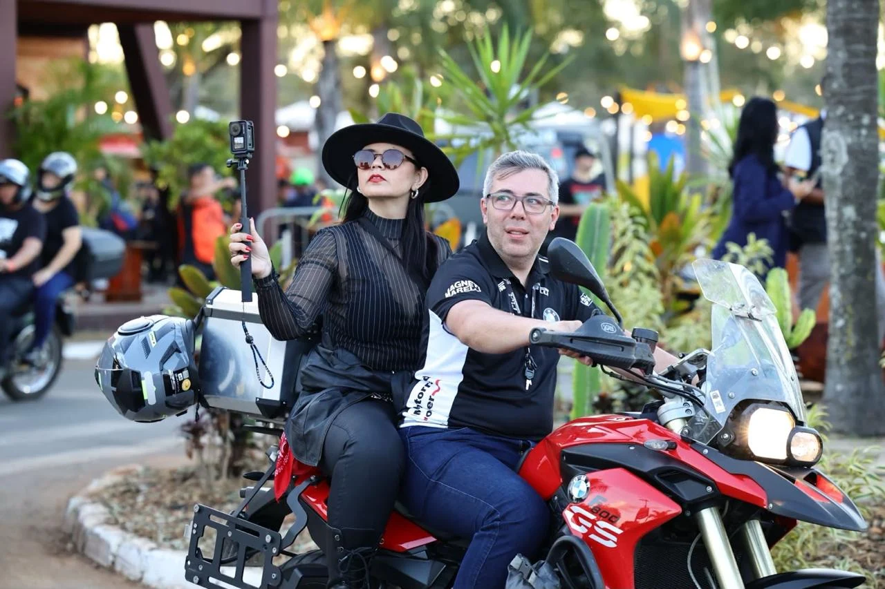 Carol de chapéu e óculos escuros na garupa de uma motocicleta durante um evento.