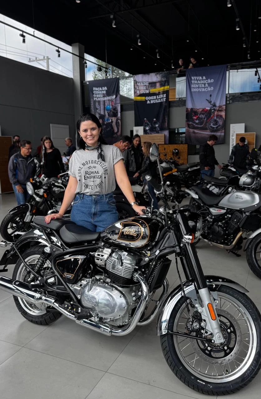 Carol ao lado de uma Royal Enfield Classic cromada em uma exposição de motocicletas.