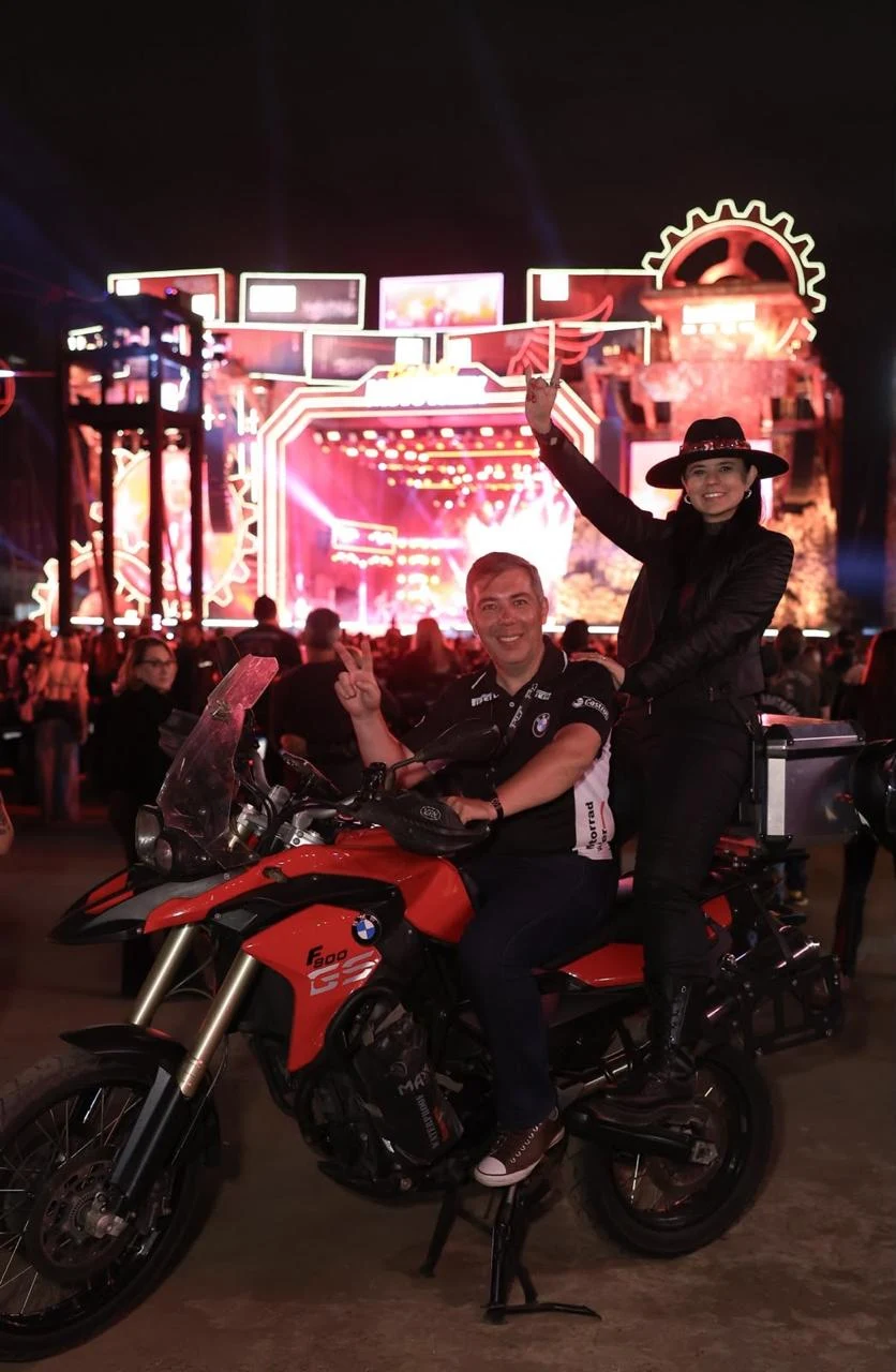 Carol em uma motocicleta diante de um palco iluminado à noite.