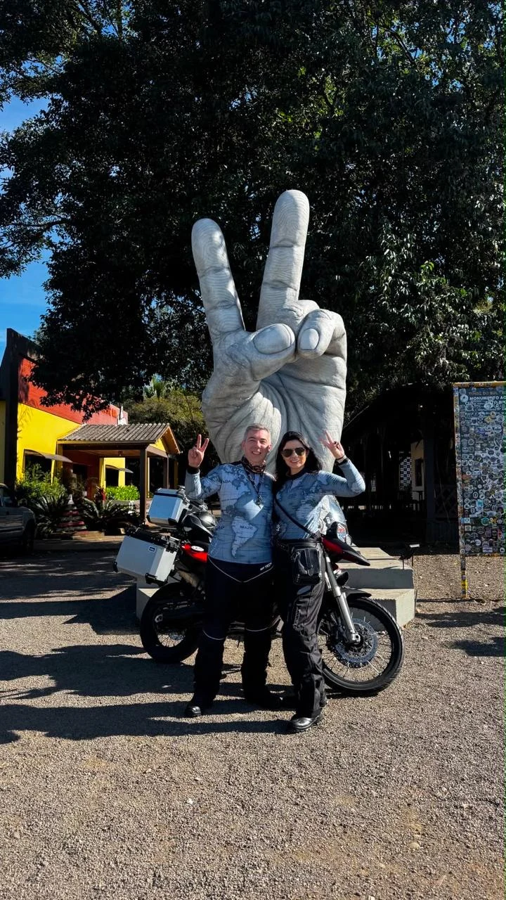 Carol e seu acompanhante ao lado da motocicleta em uma parada de viagem.