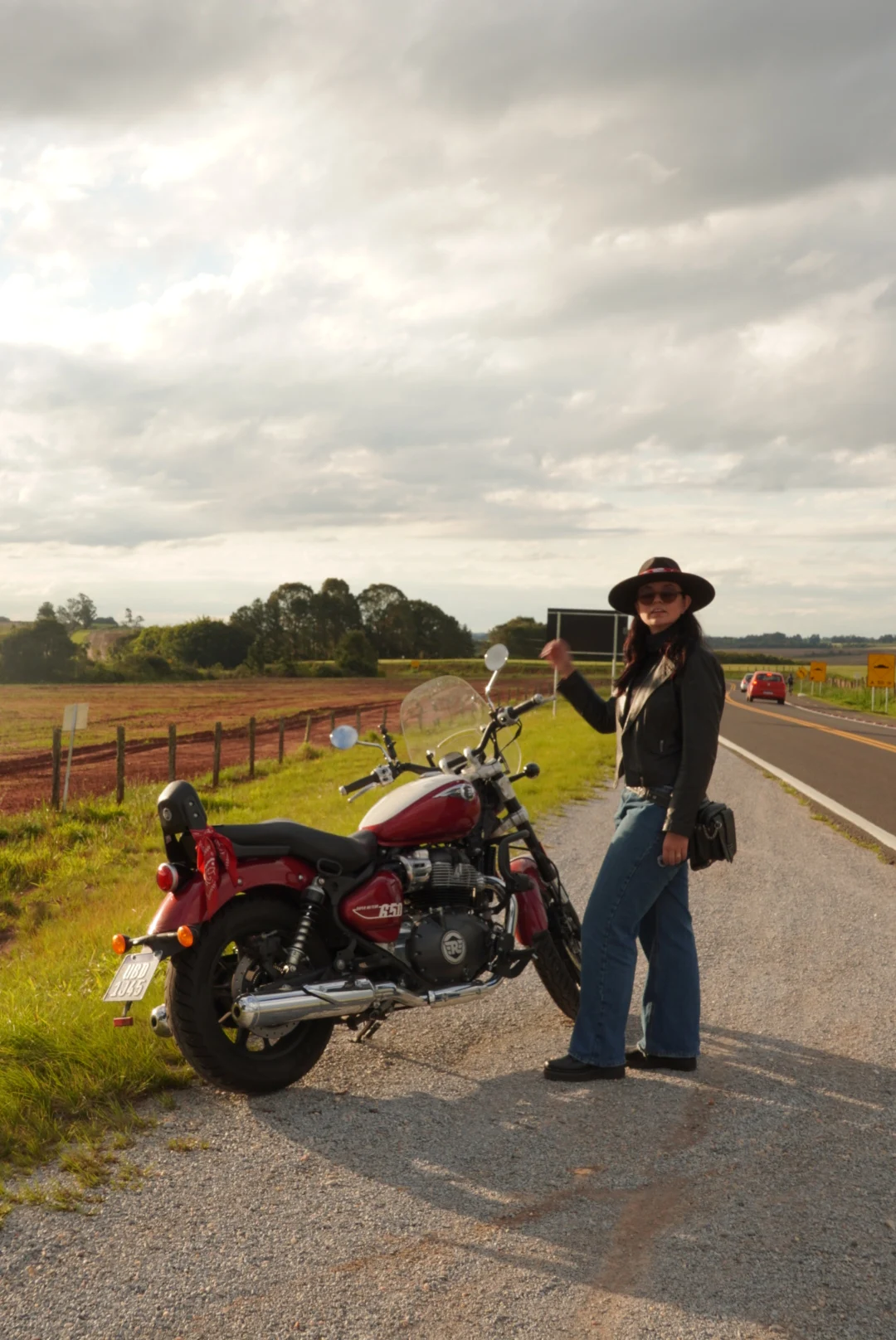 Carol Chagas de chapéu ao lado de sua motocicleta bordô na estrada.